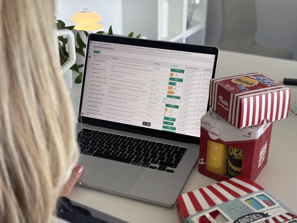 A laptop screen showing Bombiix Operational Critical Paths for a fashion and lifestyle brand. Some homeware products are next to the laptop on the desk.