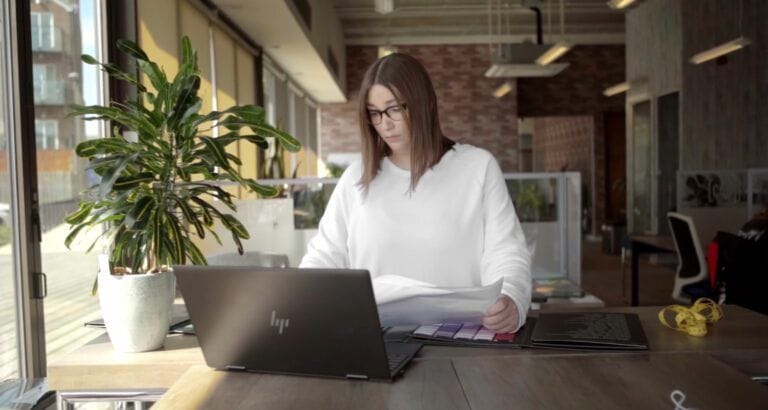 Lucy Blackley in office working on her PLM software. Her laptop is in front of her and she is reading through product designs and information. She wears a white tshirt and has brown hair and glasses.