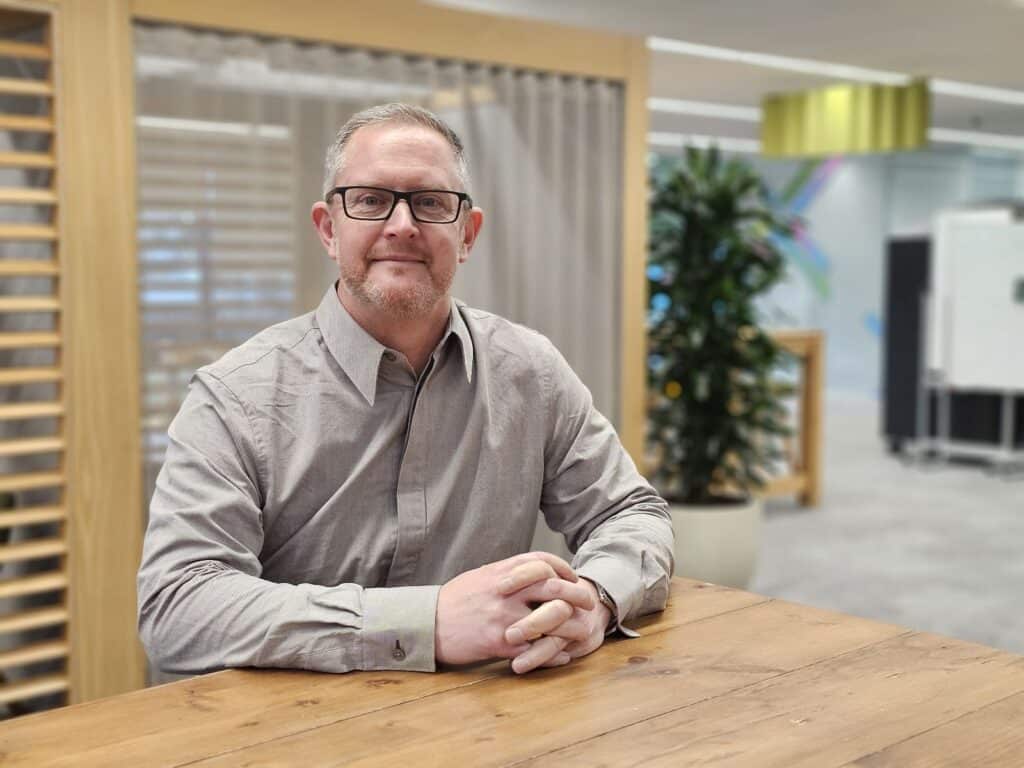 Neil Weaver, CEO of Bombiix. He sits with his hands together at a wooden desk and has a light grey shirt and glasses on.
