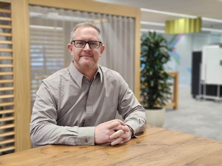 Neil Weaver, CEO of Bombiix. He sits with his hands together at a wooden desk and has a light grey shirt and glasses on.