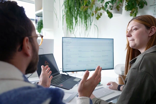 Two professionals discussing in front of a computer screen displaying a PLM collaboration dashboard.