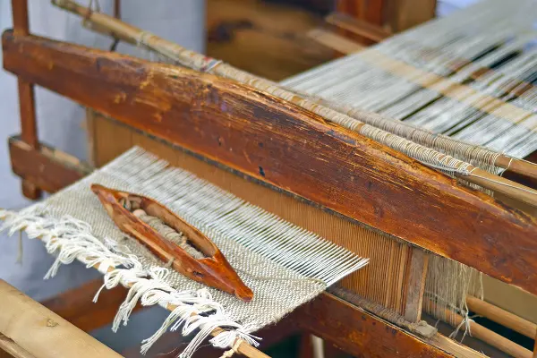 Close-up of a traditional wooden loom weaving light-coloured textile fabric.