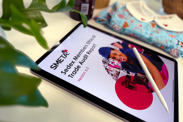 Digital tablet displaying SMETA Ethical Trade Audit Report with a stylus pen on a desk beside a baby garment.