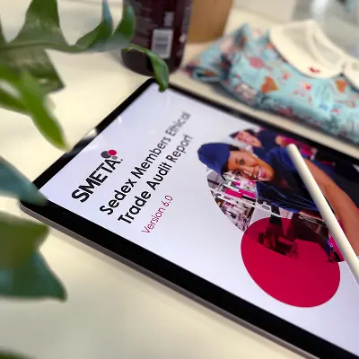 Digital tablet displaying SMETA Ethical Trade Audit Report with a stylus pen on a desk beside a baby garment.