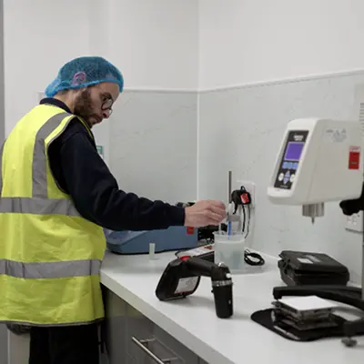 Worker in a high-visibility vest and hair net conducting product testing in a laboratory environment.