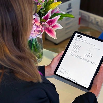 Person viewing a compliance document on a tablet at a desk with pink flowers in a vase.