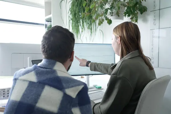 Two colleagues working together at a desk, with one pointing at a computer screen in a bright office.