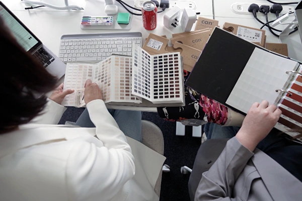 Two people reviewing fabric and colour swatch books at a desk with a keyboard, laptop, and packaging samples.