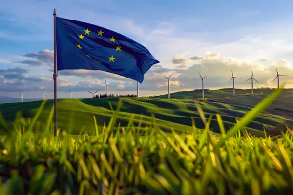 European Union flag waving in a grassy field with wind turbines in the background under a partly cloudy sky.