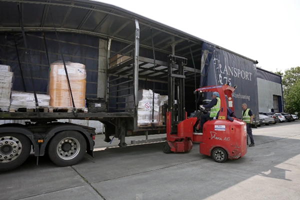 Forklift loading pallets of goods onto a large transport truck at an industrial warehouse site.