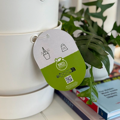Sustainability product tag showing recycling icons and “100% recycled” label attached to a white container beside a plant.