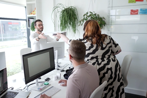 Office team collaborating in a bright workspace, with one person handing documents to a standing colleague.