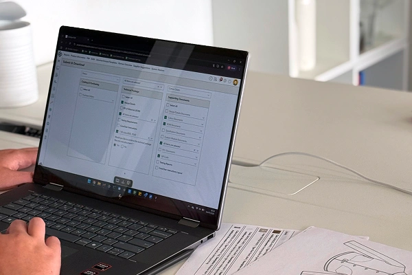 Person using a laptop displaying a tech pack management interface with printed technical drawings on the desk.