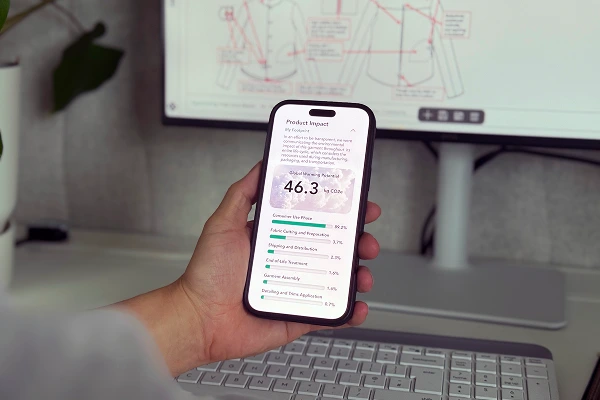 Hand holding smartphone displaying product impact and carbon footprint data, with garment technical drawings on a computer screen in the background.