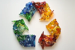 Recycling symbol created from colourful crushed plastic bottles arranged in green, blue, red, and yellow sections.
